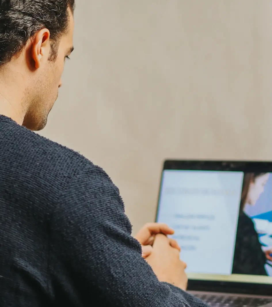 Persona con un suéter oscuro sentada en una mesa, mirando una pantalla de computadora portátil con una diapositiva de presentación borrosa visible.