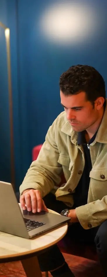 Una persona con una chaqueta beige está concentrada en el uso de una computadora portátil en una habitación con paredes de color azul oscuro.